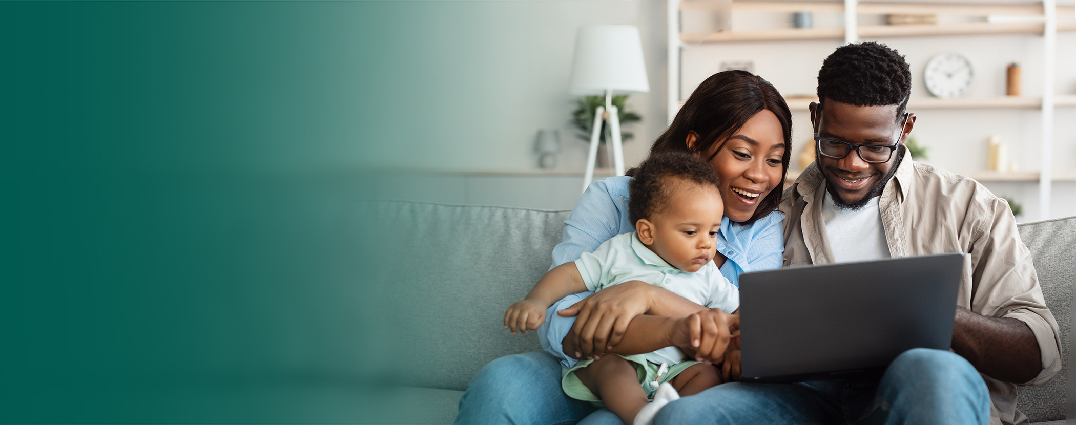 Black couple sitting on the couch with their baby, all looking at laptop screen and smiling.